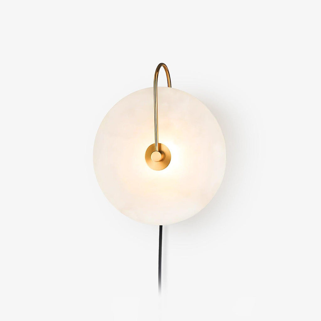 Studio shot of the Clérachic Plug-In Wall Sconce on white, gold arm lit and a black cord trailing from the alabaster disc.