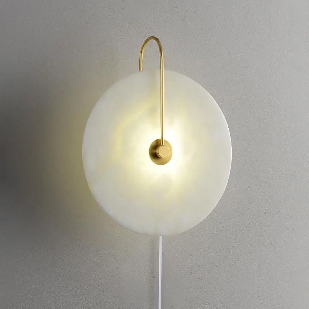 The Clérachic Plug-In Wall Sconce with a gold arm and white cord option, alabaster globe shade lit on a grey wall.