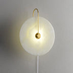 Clérachic Plug-In Wall Sconce glowing softly, white cord running down the grey plaster from the cloudy alabaster disc.