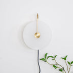 The Clérachic Plug-In Wall Sconce switched off, showing the matte alabaster globe shade and brushed gold arm by a leafy branch.