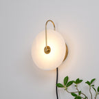Angled view of the lit Clérachic Plug-In Wall Sconce, gold mounting plate behind the alabaster disc and a plant at the edge.