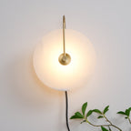 Clérachic Plug-In Wall Sconce lit head-on beside a trailing green plant, warm glow centred on the alabaster globe shade.
