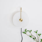 Unlit Clérachic Plug-In Wall Sconce on a white wall beside a leafy branch, gold arm hooked over the stone disc.