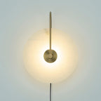 Clérachic Plug-In Wall Sconce lit against a pale backdrop, the long gold arm reaching over the alabaster globe shade.