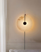 The Clérachic Plug-In Wall Sconce in black, lit on a paneled wall by a sheer curtain, casting a wide halo behind the disc.