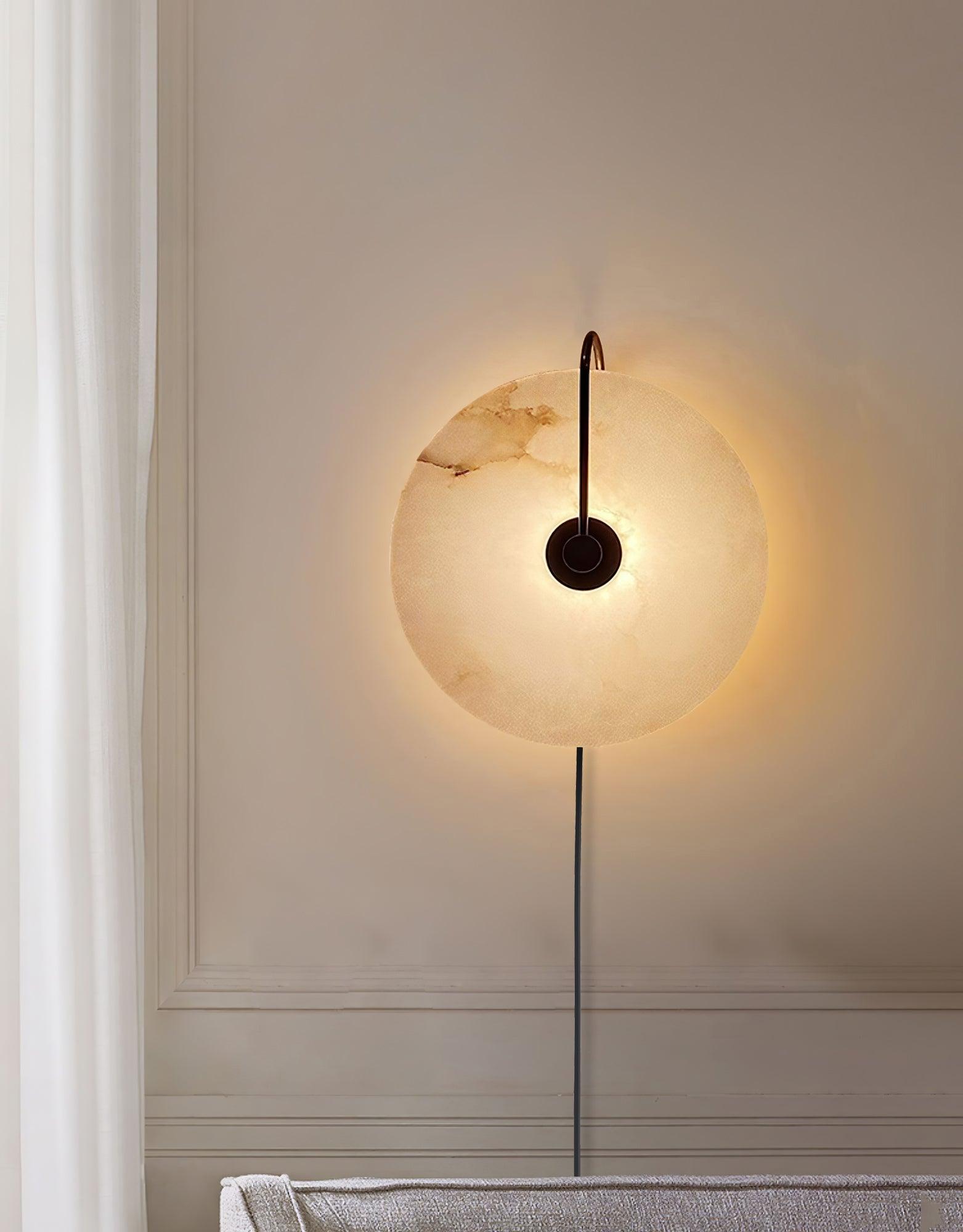 The Clérachic Plug-In Wall Sconce in black, lit on a paneled wall by a sheer curtain, casting a wide halo behind the disc.