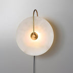 Clérachic Plug-In Wall Sconce lit on a grey wall, polished gold arm curving over the glowing alabaster globe shade.