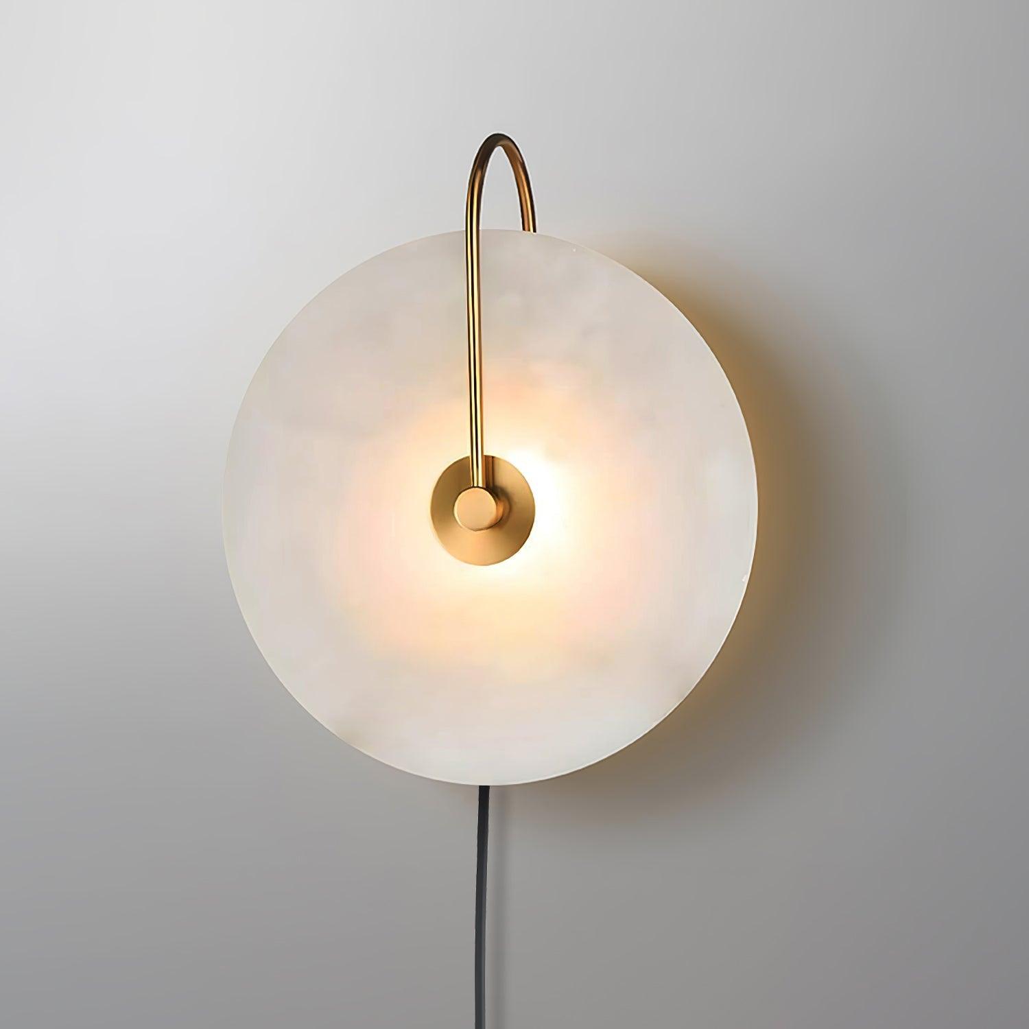 Clérachic Plug-In Wall Sconce lit on a grey wall, polished gold arm curving over the glowing alabaster globe shade.