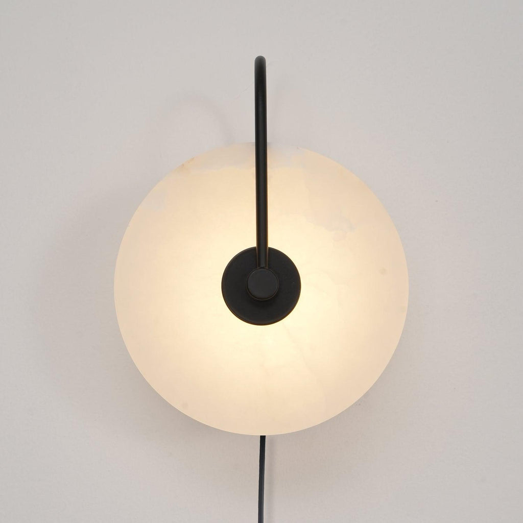 Black-arm Clérachic Plug-In Wall Sconce lit on an off-white wall, matte hardware crossing the mottled alabaster disc.