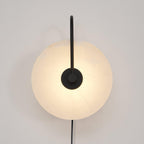 Black-arm Clérachic Plug-In Wall Sconce lit on an off-white wall, matte hardware crossing the mottled alabaster disc.
