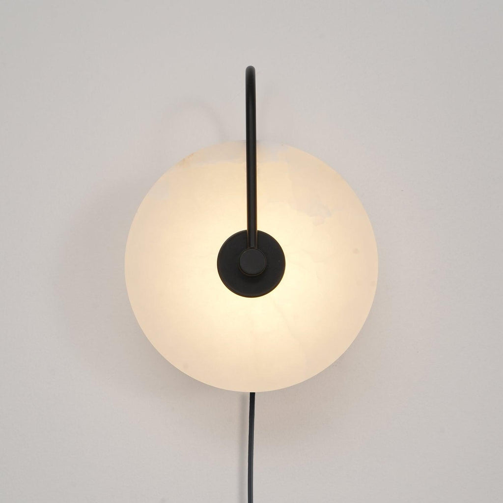 The Clérachic Plug-In Wall Sconce in black glows on an off-white wall, warm light spreading past the circular stone shade.