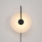 The Clérachic Plug-In Wall Sconce in black glows on an off-white wall, warm light spreading past the circular stone shade.