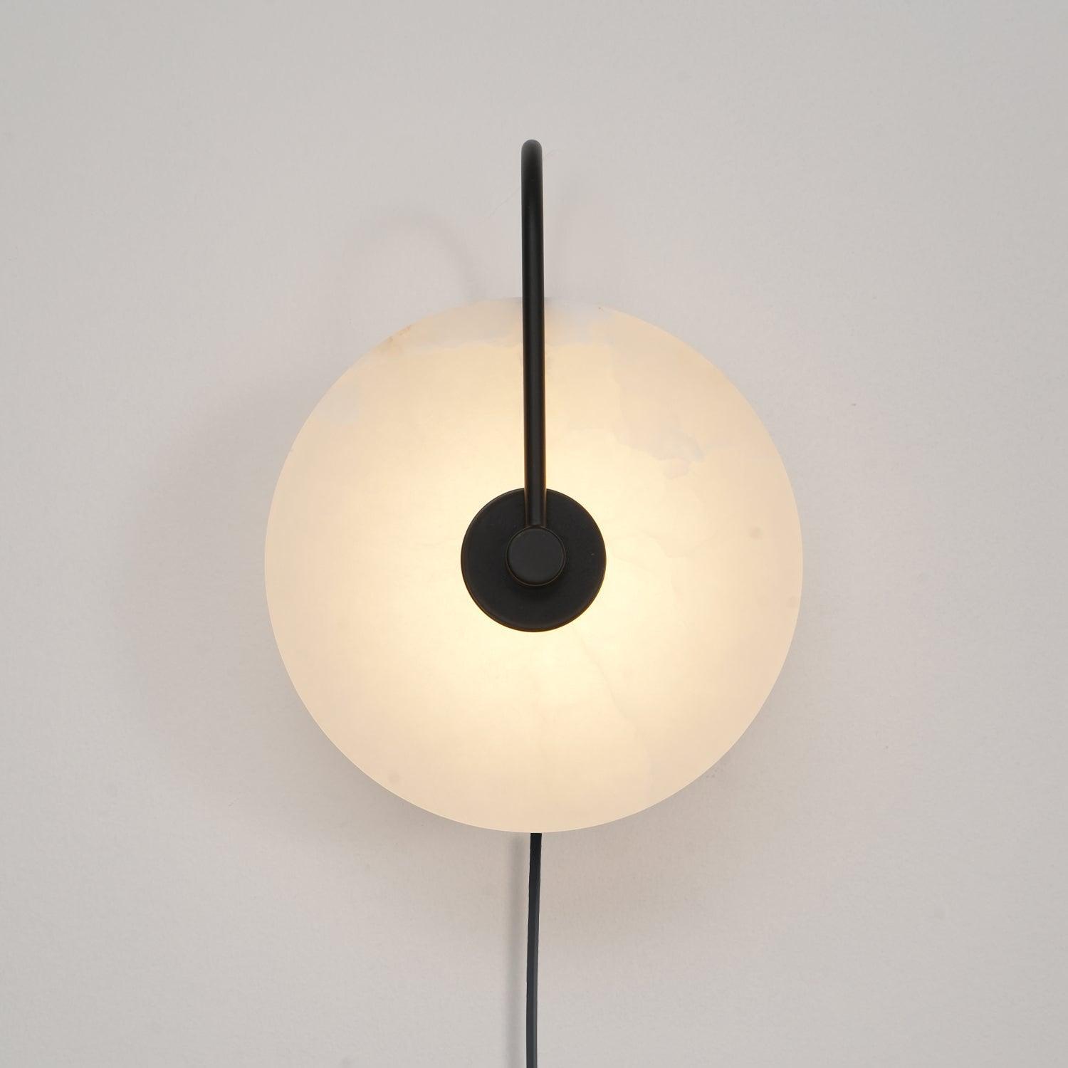 The Clérachic Plug-In Wall Sconce in black glows on an off-white wall, warm light spreading past the circular stone shade.