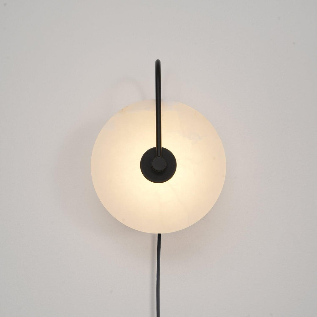 Head-on Clérachic Plug-In Wall Sconce in black on a white wall, slim vertical arm and grey veining in the lit disc.
