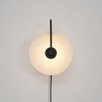 Head-on Clérachic Plug-In Wall Sconce in black on a white wall, slim vertical arm and grey veining in the lit disc.