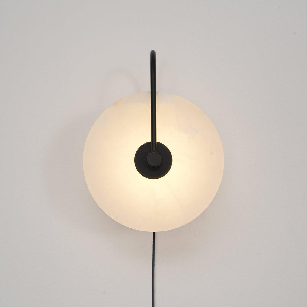 Clérachic Plug-In Wall Sconce with a matte black arm, the alabaster globe shade glowing warm on a plain off-white wall.