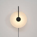 Clérachic Plug-In Wall Sconce with a matte black arm, the alabaster globe shade glowing warm on a plain off-white wall.