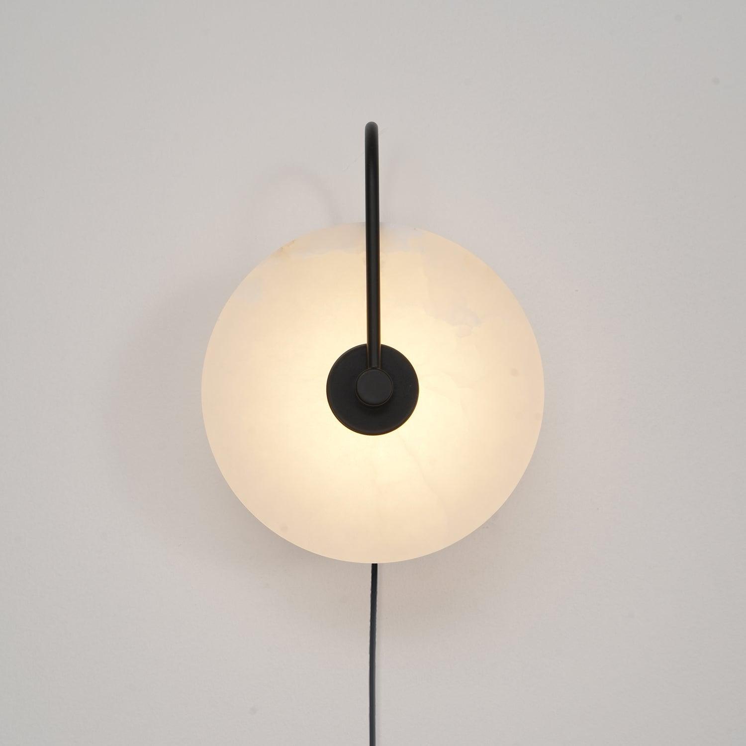 Clérachic Plug-In Wall Sconce with a matte black arm, the alabaster globe shade glowing warm on a plain off-white wall.