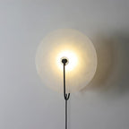Lit Clérachic Plug-In Wall Sconce in black on a light grey wall, forked metal arm holding the glowing stone disc.