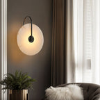 Clérachic Plug-In Wall Sconce in black on a dark wall, alabaster disc lit above a fern in a gold planter and a tufted chair.