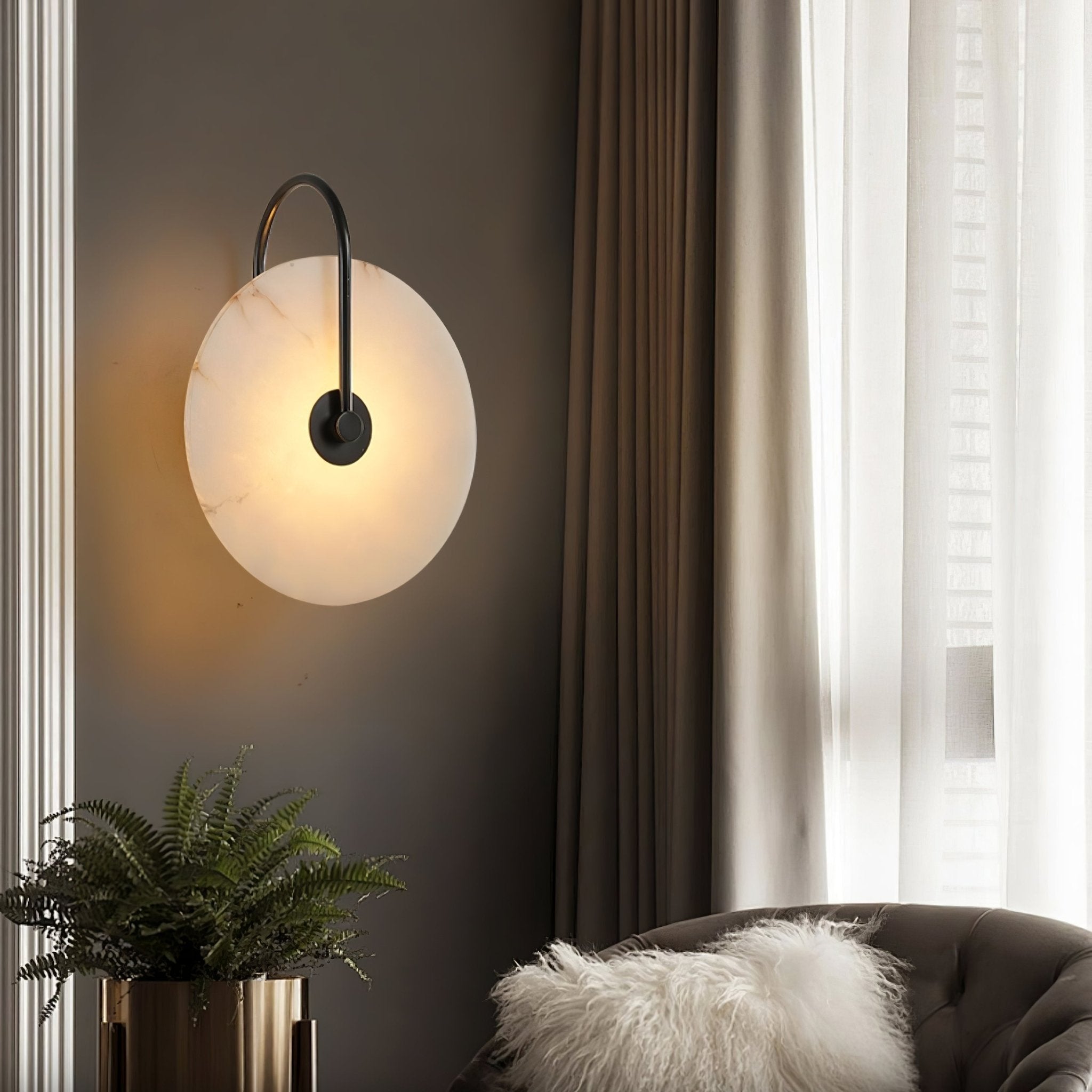Clérachic Plug-In Wall Sconce in black on a dark wall, alabaster disc lit above a fern in a gold planter and a tufted chair.