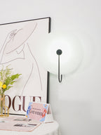 Lit Clérachic Plug-In Wall Sconce in black on a white wall, beside a line-drawing fashion poster and yellow flowers.