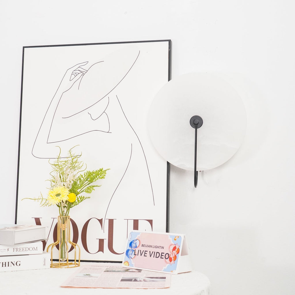 Unlit Clérachic Plug-In Wall Sconce in black beside a fashion poster, gold wire vase and a yellow daisy on the table.