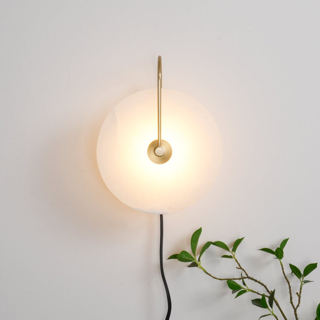 Clérachic Plug-In Wall Sconce glowing on an off-white wall, gold arm and black cord with a plant tip at the lower edge.