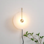 Clérachic Plug-In Wall Sconce glowing on an off-white wall, gold arm and black cord with a plant tip at the lower edge.