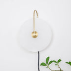 Unlit Clérachic Plug-In Wall Sconce on an off-white wall, gold arm over the pale disc and a green plant sprig at the corner.