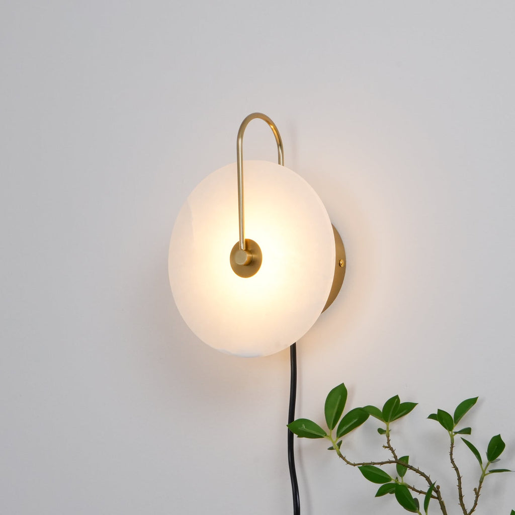 Clérachic Plug-In Wall Sconce glowing on a white wall, gold hook arm holding the warm alabaster globe shade in place.