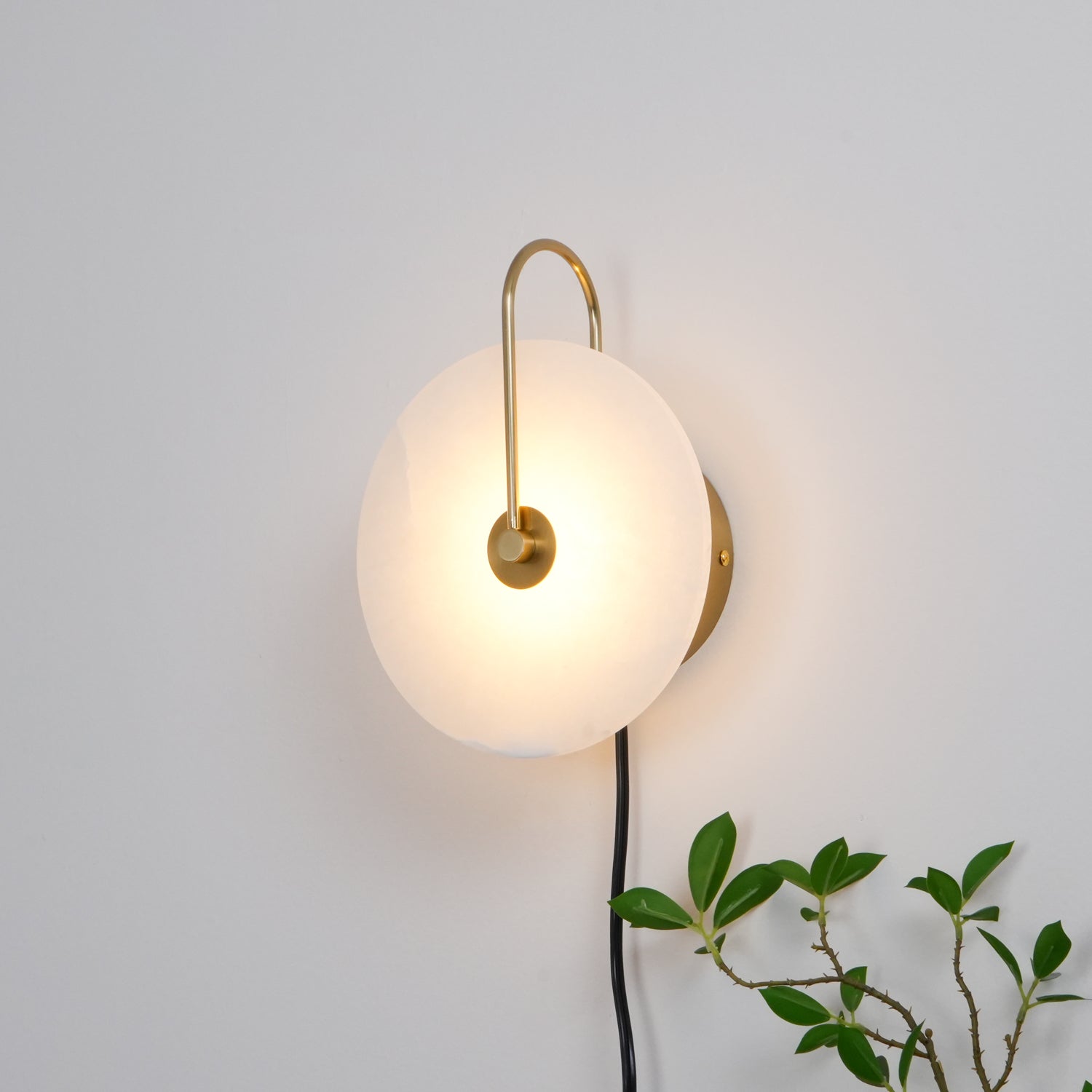 Clérachic Plug-In Wall Sconce glowing on a white wall, gold hook arm holding the warm alabaster globe shade in place.