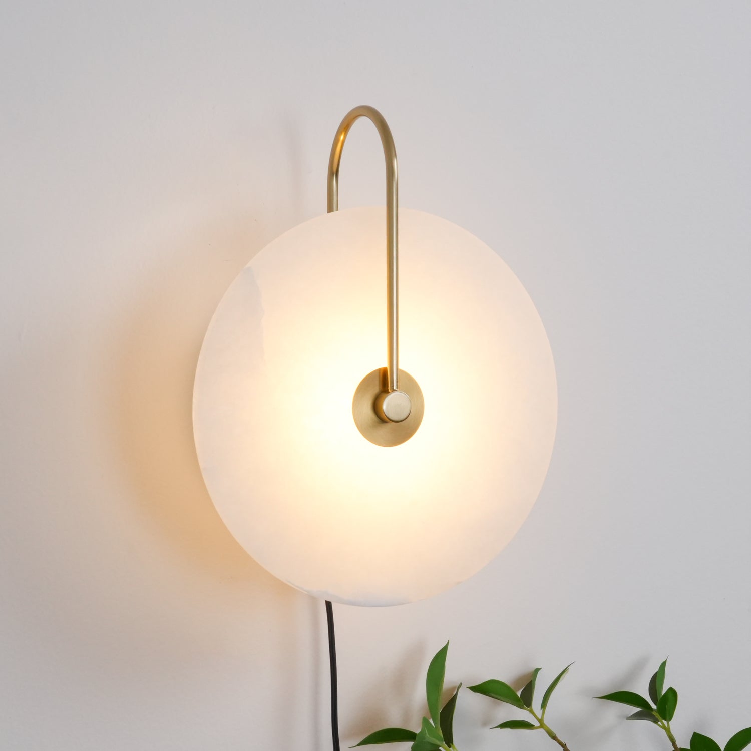 Clérachic Plug-In Wall Sconce on a light grey wall, gold U-shaped arm and glowing disc with leafy sprigs at the corner.