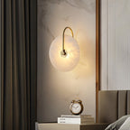 Clérachic Plug-In Wall Sconce on a marble bedside panel, gold arm glowing above stacked books and a twin-bell clock.