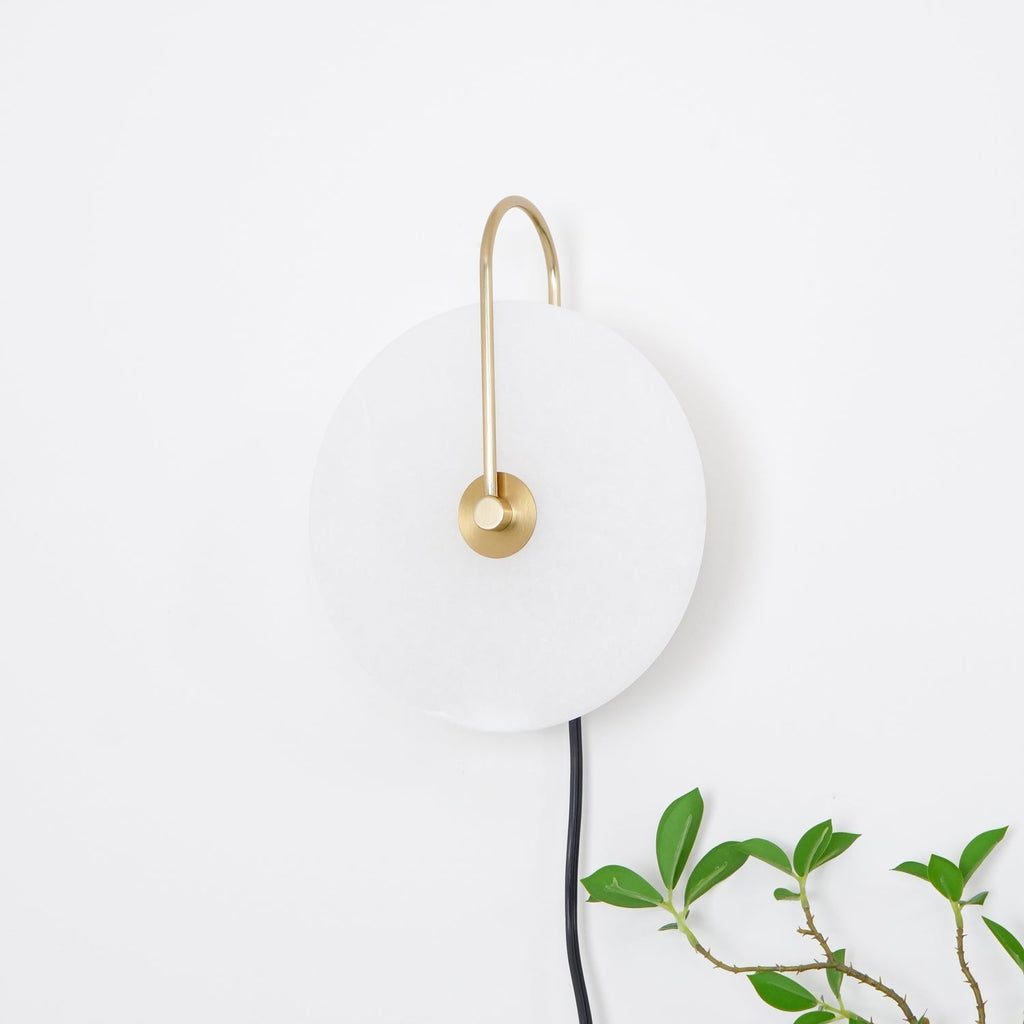 Clérachic Plug-In Wall Sconce on a white wall, satin gold arm and black cord beside a leafy plant with brown stems.