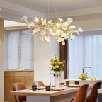 Lit Clérachic chandelier of white ginkgo leaves over a kitchen island with tan leather stools, with yellow flowers and sheer curtains behind.