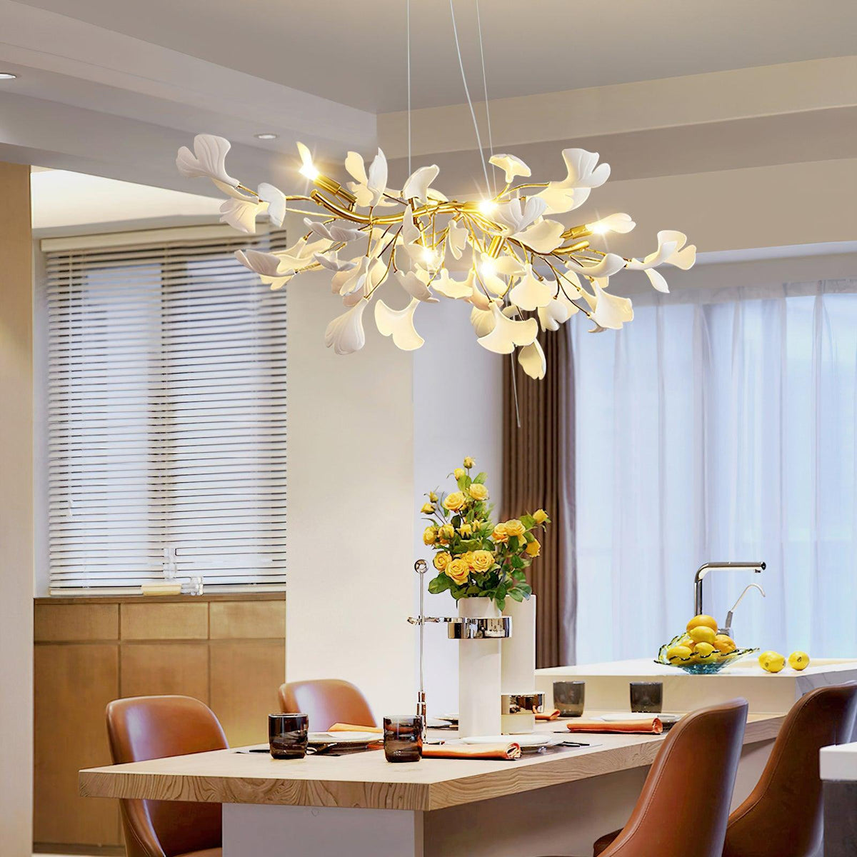 Lit Clérachic chandelier of white ginkgo leaves over a kitchen island with tan leather stools, with yellow flowers and sheer curtains behind.