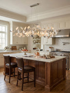 Long Clérachic leaf chandelier suspended over a walnut kitchen island with leather bar stools, in a cream kitchen with white cabinetry.