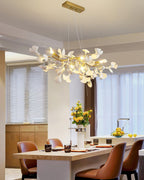 White porcelain leaves and gold branches of this Clérachic chandelier, lit above a pale kitchen counter with caramel leather chairs.