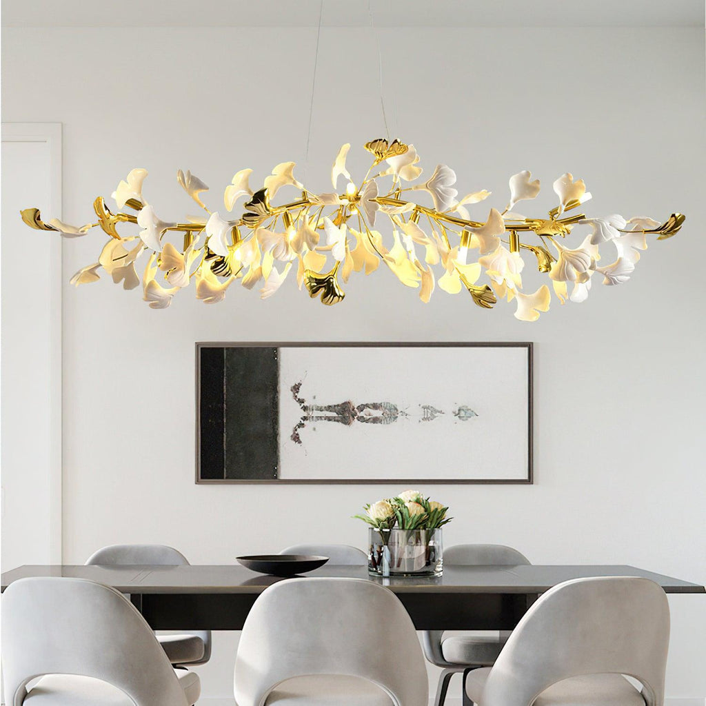 Wide Clérachic chandelier with gold branch tips and lit white leaves above a black dining table, with grey chairs and a framed print behind.