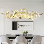 Wide Clérachic chandelier with gold branch tips and lit white leaves above a black dining table, with grey chairs and a framed print behind.