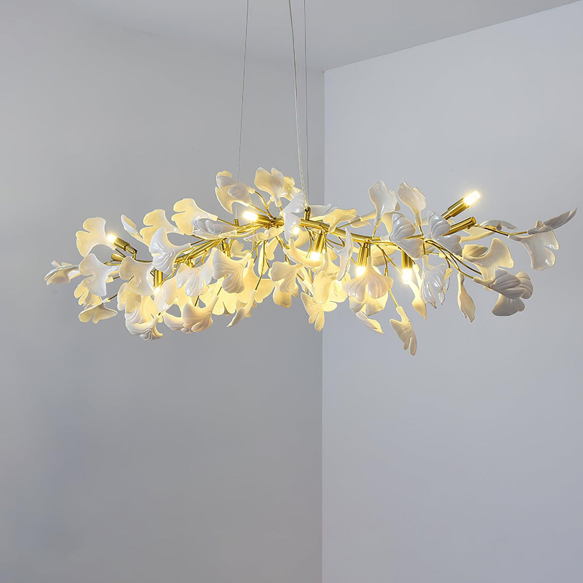 Lit Clérachic chandelier of white ginkgo leaves and small candle-style bulbs, hung in a room corner with pale grey walls.