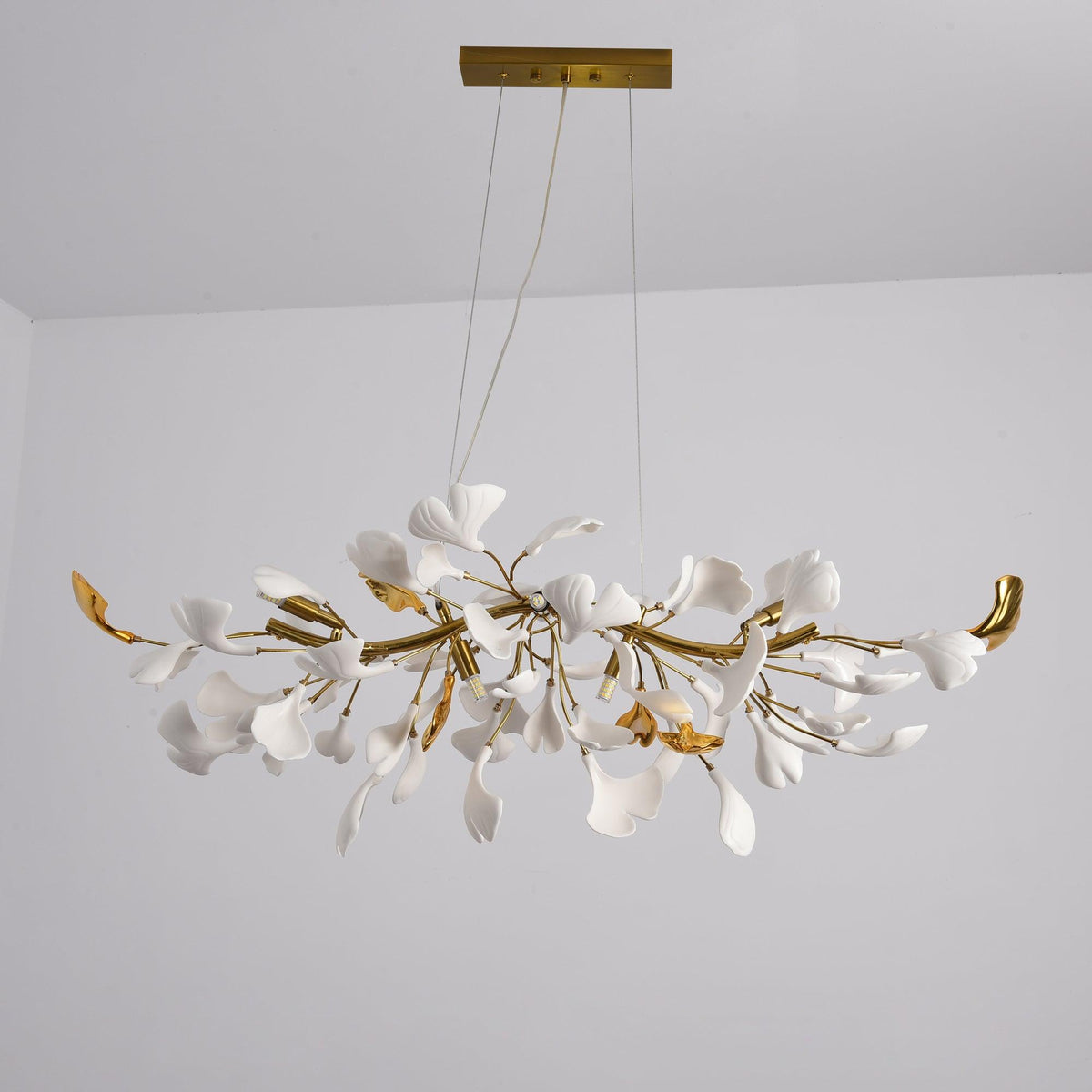 Unlit Clérachic chandelier, white leaves among gold branches with curved gold tips, hanging from two cables under a gold canopy.