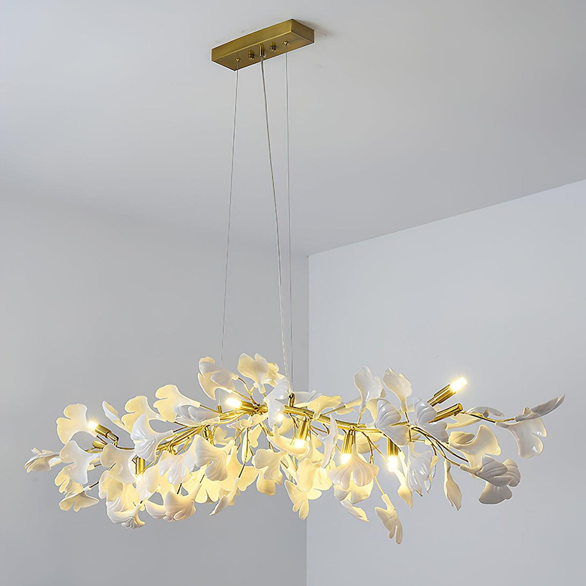 Clérachic leaf chandelier hanging from a gold canopy in a pale grey corner, bulbs on and white leaves lit from within.