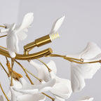 Close view of a gold lamp arm with an LED capsule among white porcelain leaves on this Clérachic chandelier, against a grey ceiling.