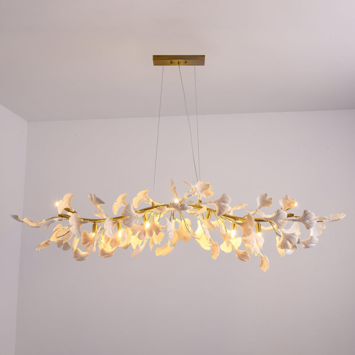 Wide Clérachic chandelier lit and hung low in a white-walled room, its white leaves glowing warm along a gold branch.