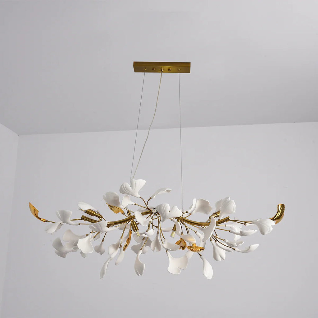 Clérachic chandelier with gold branches and white leaves, photographed from slightly below against a pale grey wall and ceiling.