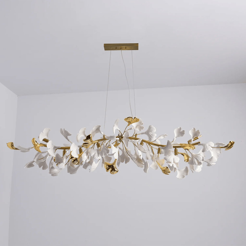 Unlit Clérachic chandelier with gold curved branches and white leaves, hanging from a gold canopy in a grey room corner.