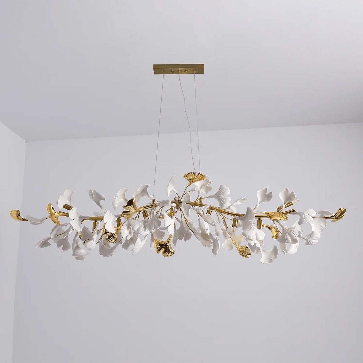Unlit Clérachic chandelier with gold curved branches and white leaves, hanging from a gold canopy in a grey room corner.