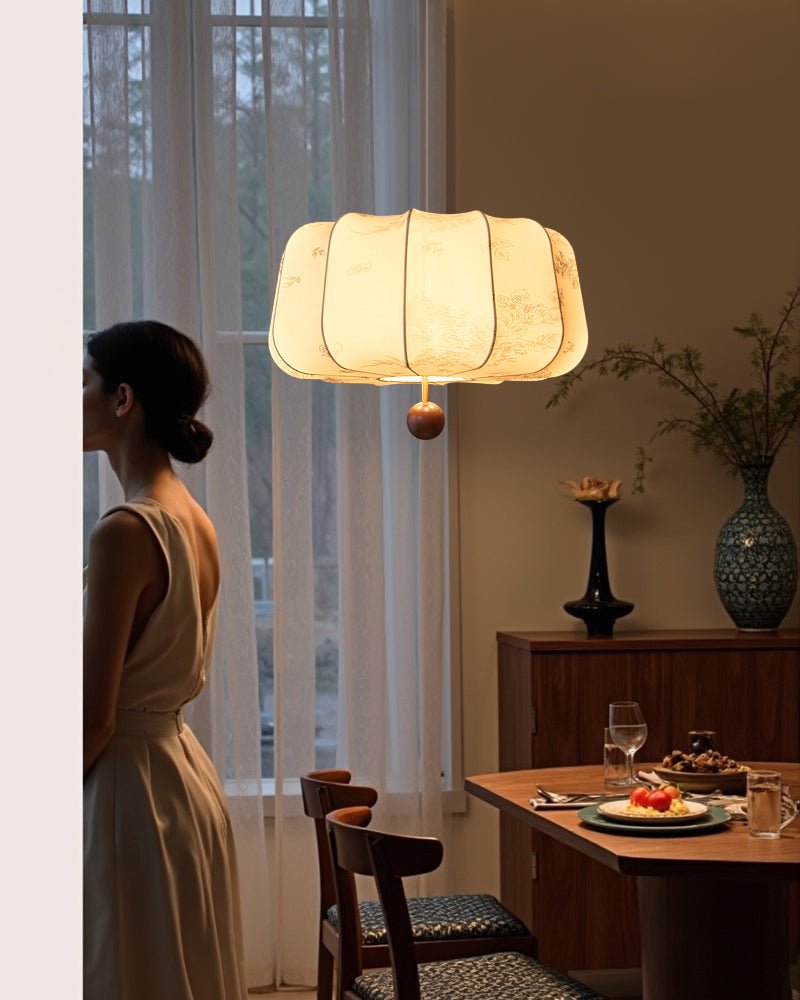 Clérachic scalloped fabric pendant light seen in profile, lit above a dining table set with a wine glass, as a woman looks toward sheer curtains.